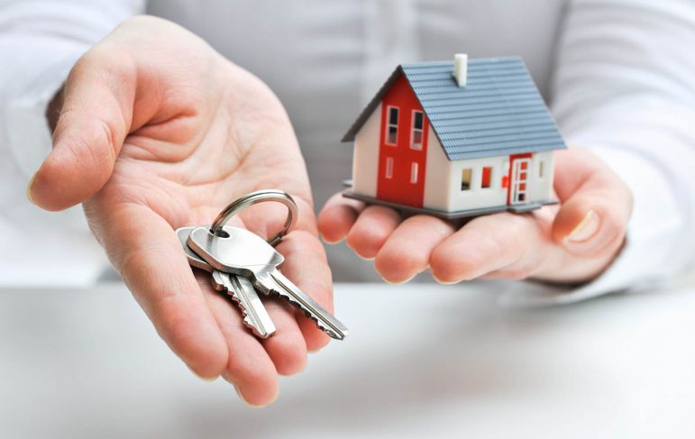 A person holds a key in one hand and a small house model in the other, symbolizing home ownership and new beginnings.