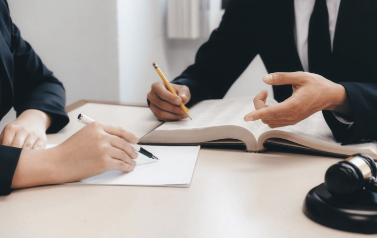 Two professionals in business attire seated at a desk, engaged in discussion with a pen and paper in front of them.